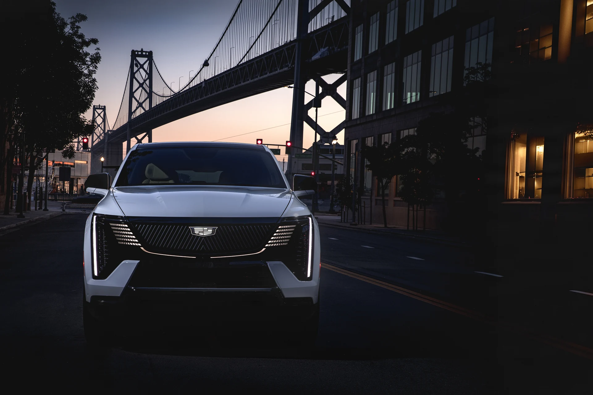 Белый Cadillac Escalade IQ 2025 едет по улице Сан-Франциско под мостом Бэй-Бридж в сумерках, почти анфас